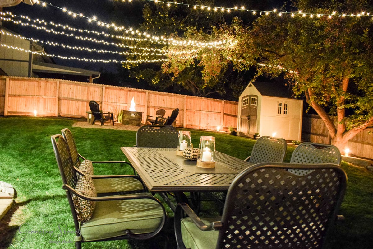 A backyard at night with string lights overhead, illuminating a patio set with six chairs around a table. A fire pit area with additional chairs is visible, and a small shed stands near a tree. The setting is cozy and inviting.