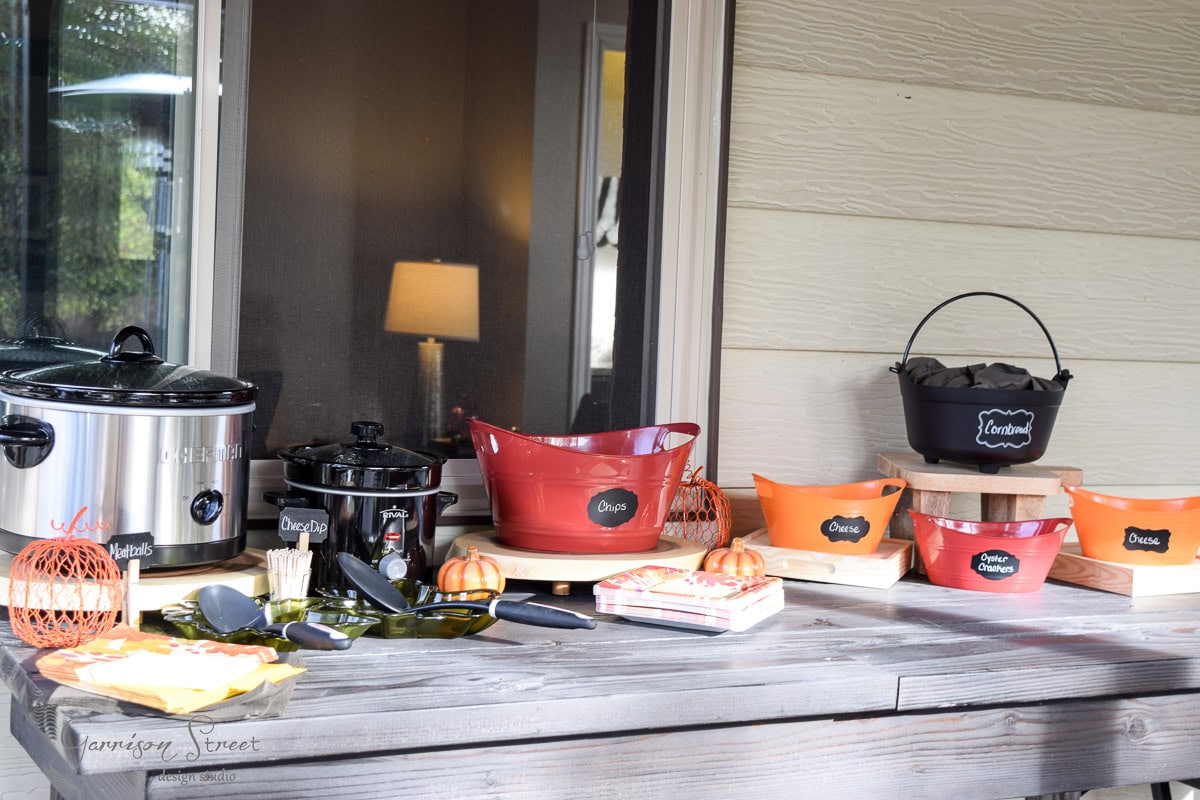 A buffet table set up outdoors, featuring slow cookers, serving baskets with dips labeled in chalk, and decorative pumpkins. There are small bags labeled "Spoons" and "Scones," along with plates and utensils arranged neatly.