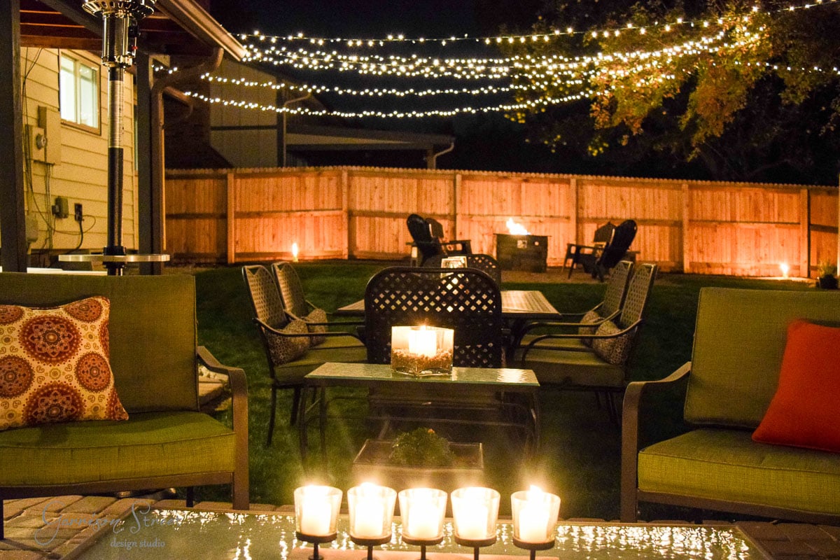 A cozy backyard at night with string lights overhead. Several candles illuminate the space, surrounded by outdoor seating and tables. The fence encloses the area, creating a warm, inviting atmosphere.