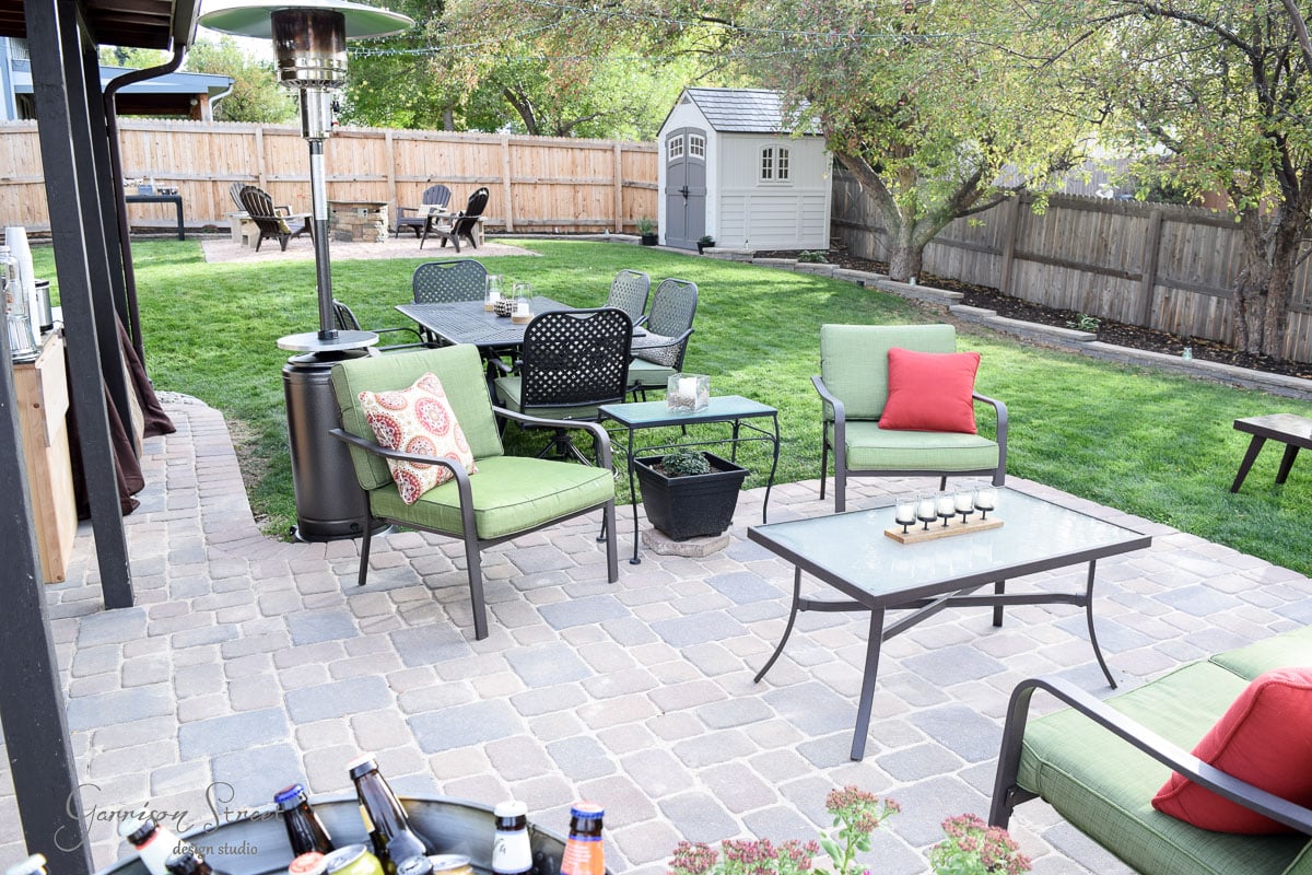 Outdoor patio with green cushioned chairs, a glass-top table, and a patio heater on a stone-paved area. A grassy lawn, trees, and a wooden fence surround the space. A small shed is in the corner, with various bottles and flowers in the foreground.