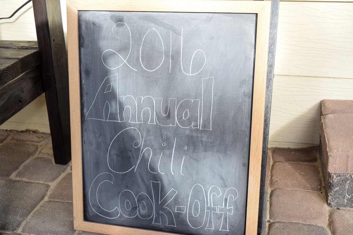 A chalkboard sign with "2016 Annual Chili Cook-Off" written on it in white chalk. The board is framed in wood and placed on a stone or brick pavement.