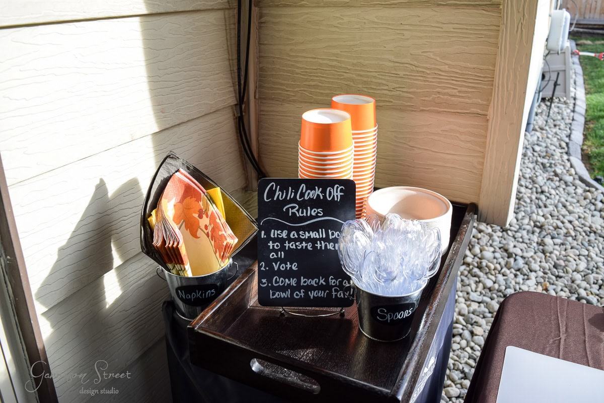 Outdoor setup for a chili cook-off with stacked orange cups, spoons in a bucket, napkins, and a chalkboard. Chalkboard lists rules: 1. Use a small cup. 2. Vote. 3. Return for more. Pebble path and part of a table visible.