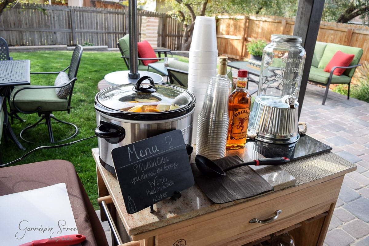 Outdoor beverage station on a patio with a chalkboard menu listing mulled cider. Items include a crockpot with citrus slices, cups, a whiskey bottle, glass dispenser, ladle, and a wooden stirrer. Patio furniture and greenery are in the background.