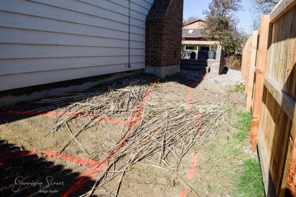 Backyard area with bare soil and scattered dried branches. Orange spray paint marks lines and shapes on the ground. A wooden fence is on the right, and a house with brick and siding is on the left. A small patio with furniture is visible in the background.