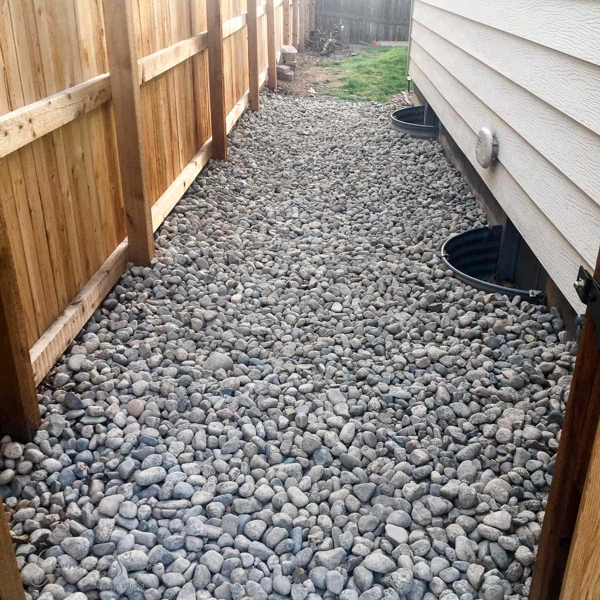 A narrow pathway covered with small gray stones runs between a wooden fence on the left and a beige-sided building on the right. Two round, black drainage covers are embedded in the stones near the building. A patch of grass is visible at the end.