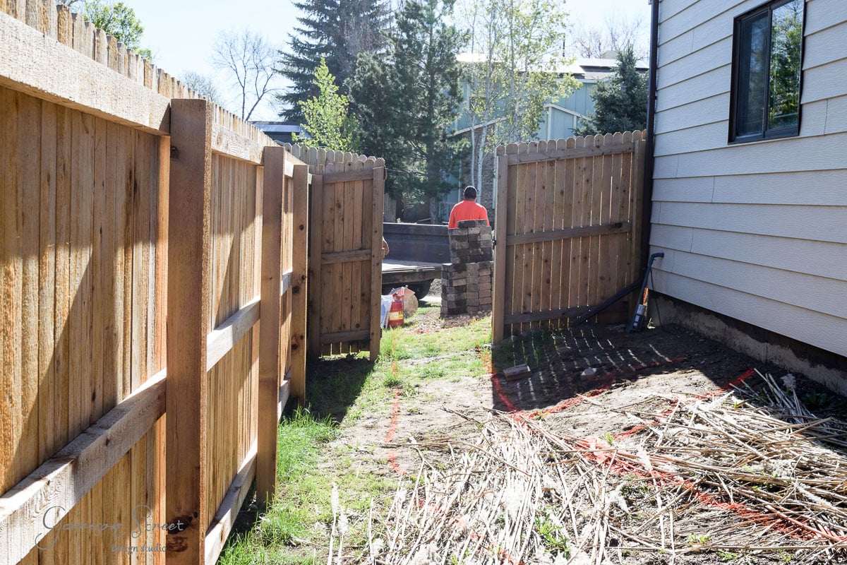 A person in a red shirt works on a backyard patio construction project, surrounded by fencing. The ground is covered with dirt and straw, and there are trees and houses in the background.