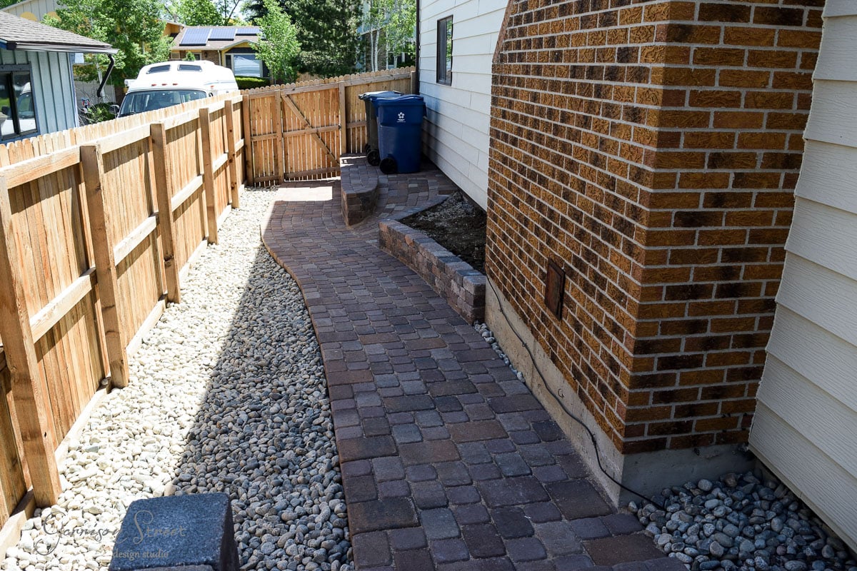 A narrow backyard features a curved stone walkway bordered by round white pebbles. A wooden fence runs alongside, and a brick house with a small garden bed and a blue recycling bin is on the right. A trailer is visible in the background.