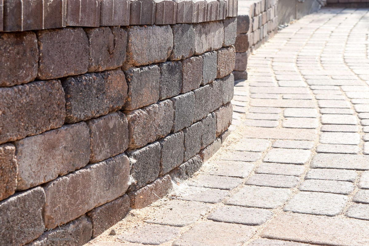 Curved stone retaining wall made of stacked bricks, adjoining a smoothly curved brick walkway. The bricks are a mix of dark gray and brown tones, and the sunlight highlights the texture of the masonrywork.