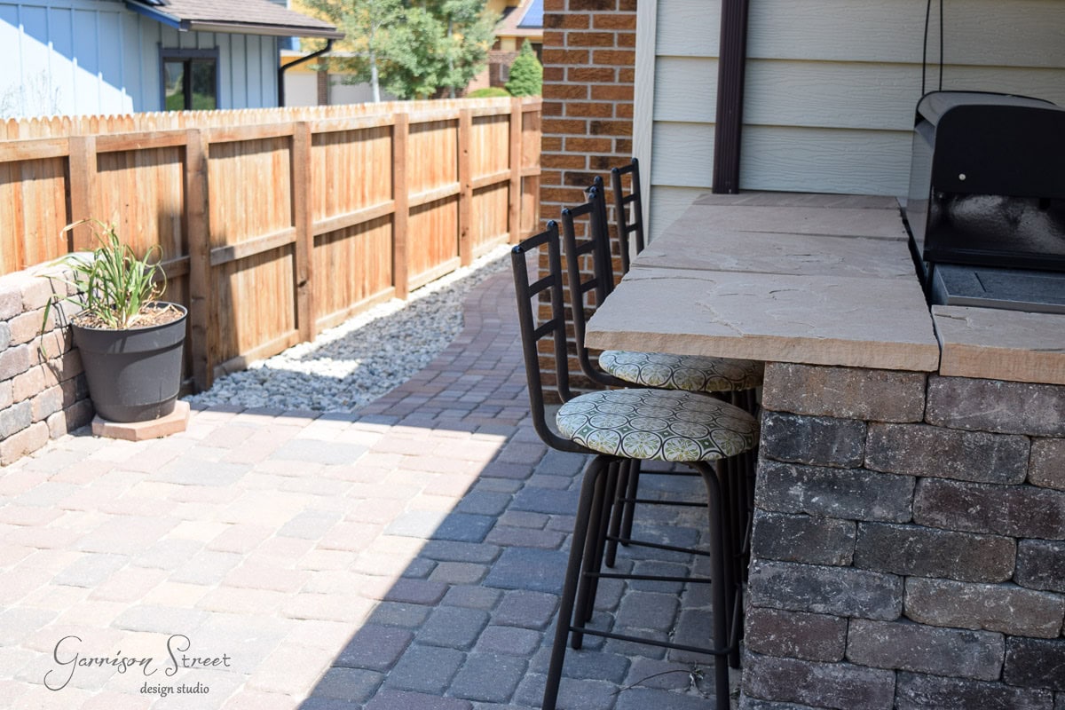 Outdoor patio with stone bar counter and three bar stools featuring patterned cushions. A small grill is on the counter. The patio has a paver floor, wooden fence, and a potted plant on a gravel path. Nearby houses and trees are visible.