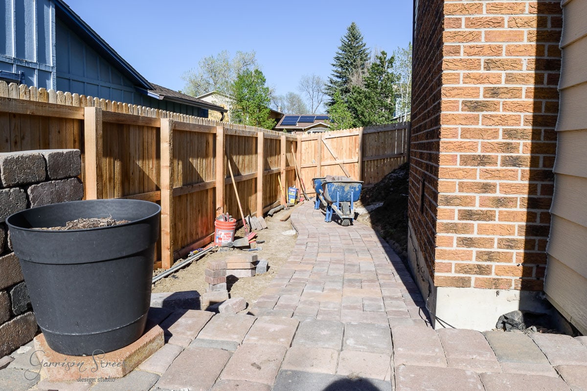 A narrow, paved walkway runs beside a brick house, leading to the backyard. A wooden fence and a block wall border the path. A large black planter and gardening tools, including a wheelbarrow and buckets, are placed along the path.