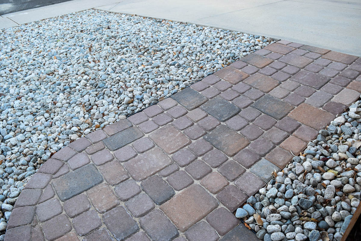 A walkway made of brown paving stones borders areas covered with small gray stones. The path curves slightly, contrasting with the light concrete sidewalk in the background.