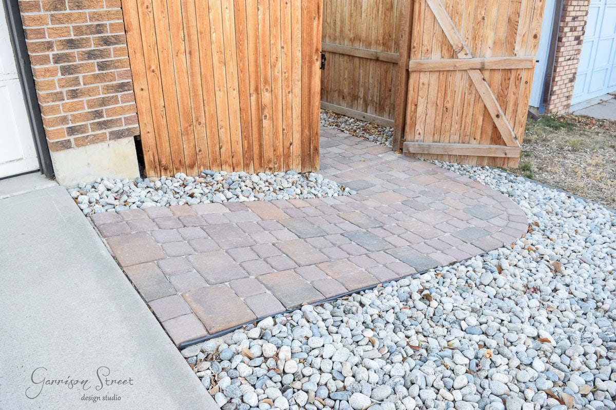 A curved stone path with interlocking pavers runs between a wooden fence and a gravel area with small stones. The fence is partially open, revealing more gravel beyond. A brick building is visible to the left.