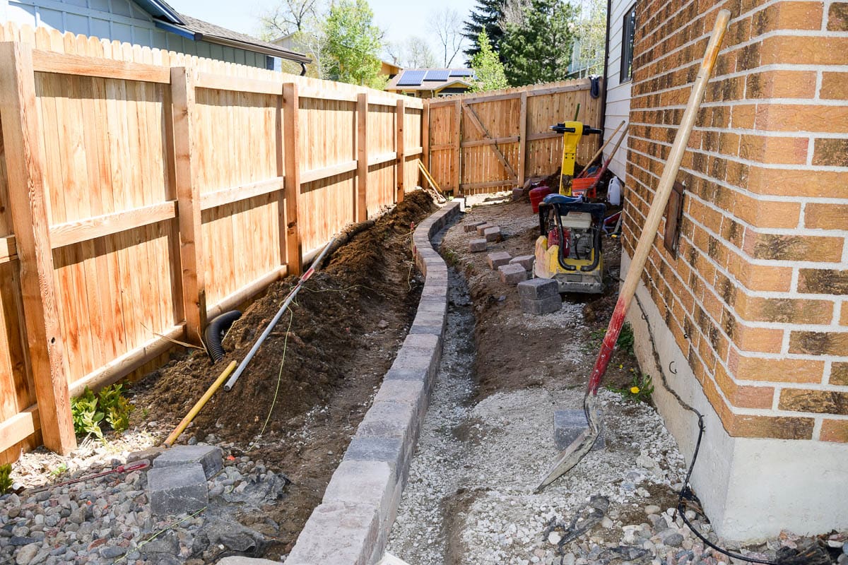 A backyard under construction with a partially built stone retaining wall alongside a wooden fence. Various construction tools, including a compactor, are present. The ground is uneven and shows exposed dirt and gravel.