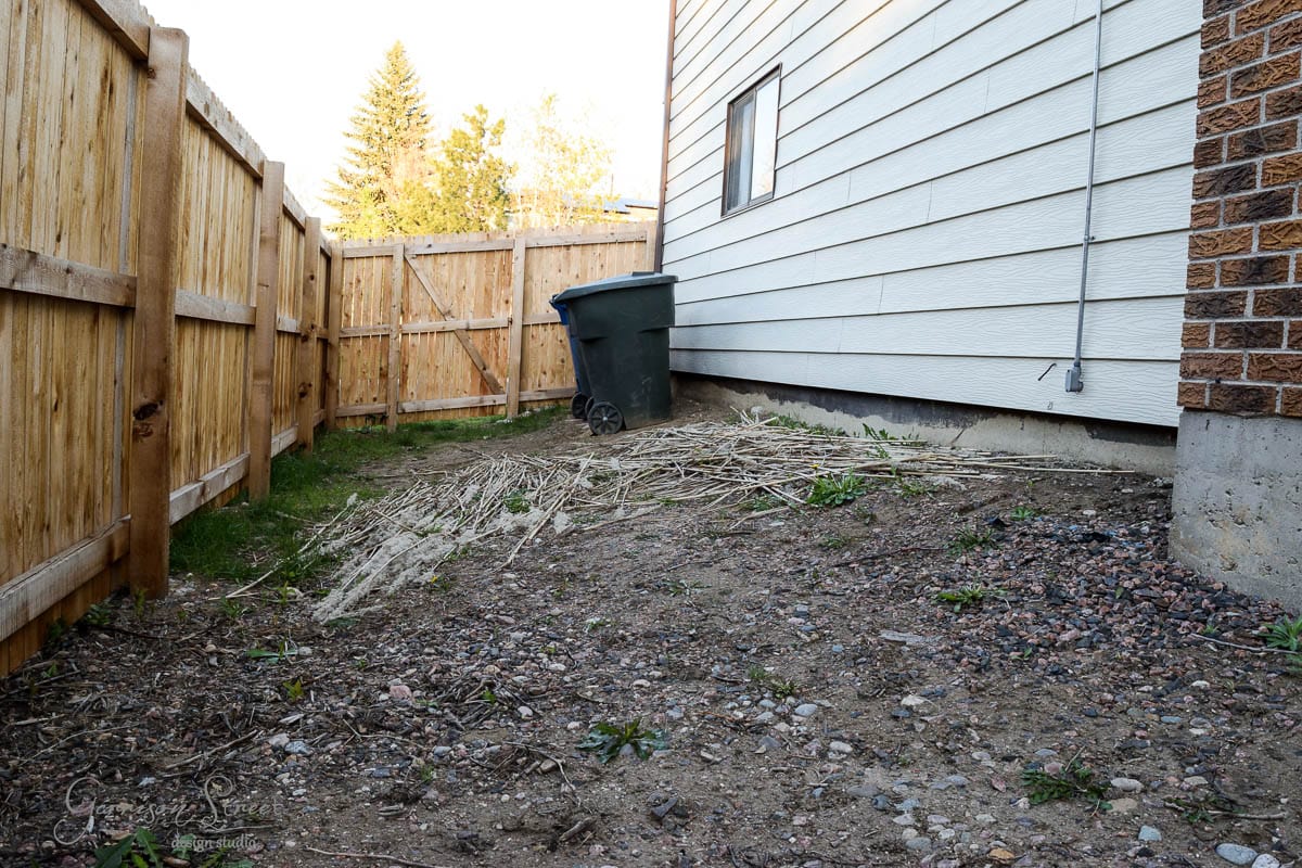 Narrow side yard with patchy dirt and scattered rocks. A wooden fence and the side of a house enclose the area. A green garbage bin is near the house. Sparse grass and weeds are visible. A small window is on the house wall. Trees are in the background.