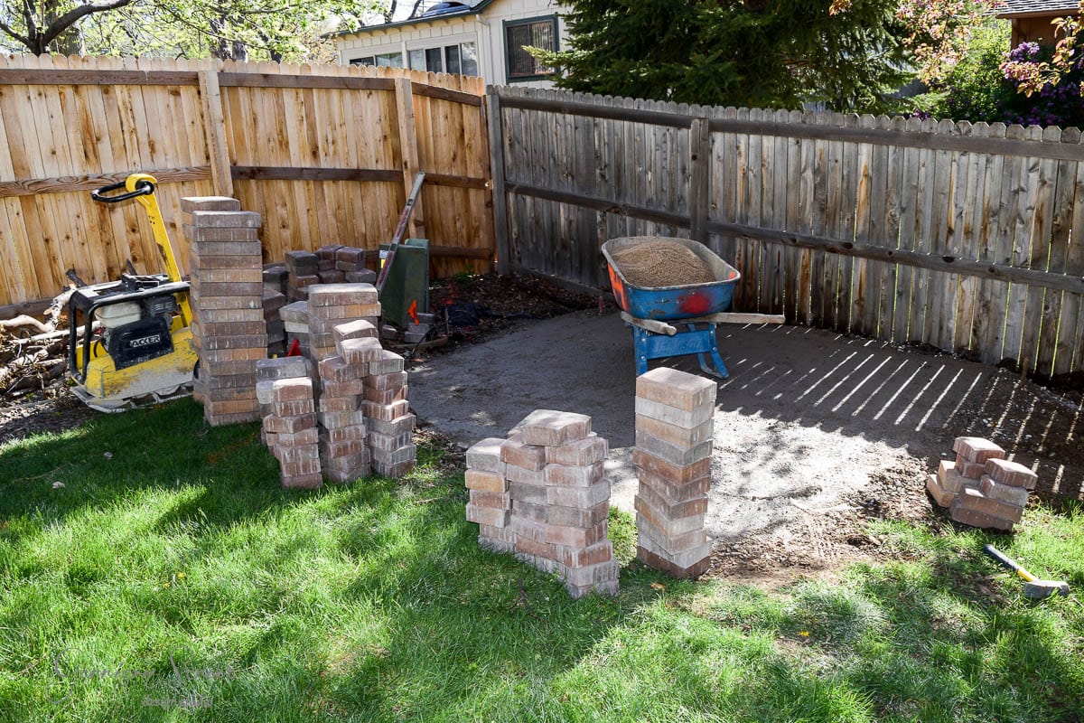 A backyard area with stacks of bricks, a red wheelbarrow holding sand, and a yellow compactor. Two wooden fences form a corner behind the workspace, with a patch of green grass in the foreground. Sunlight casts shadows across the scene.