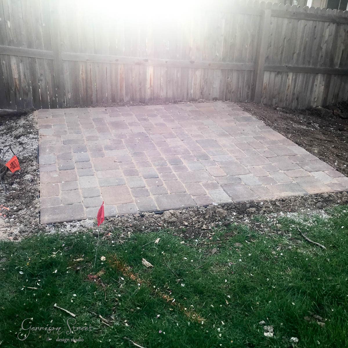 A newly constructed square patio made of stone pavers is set in a backyard, surrounded by dirt and grass. A wooden fence stands behind it, and a couple of red flags mark the area.