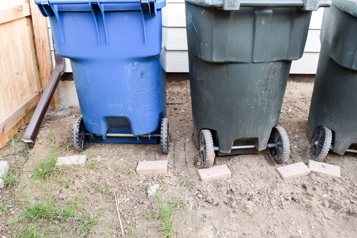Three trash bins on wheels are lined up outdoors: a blue bin on the left and two green bins on the right. They are placed on dirt, each resting on two bricks. A wooden fence and a house wall are visible in the background.