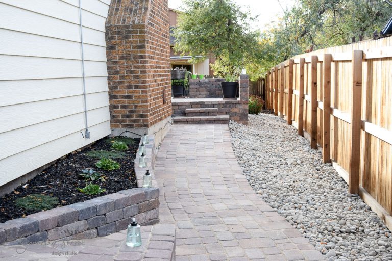 A narrow backyard features a stone path leading to a raised patio with a black planter. On the left, there's a small garden bed with plants and lanterns. A wooden fence borders the right side, with a gravel strip beside it.