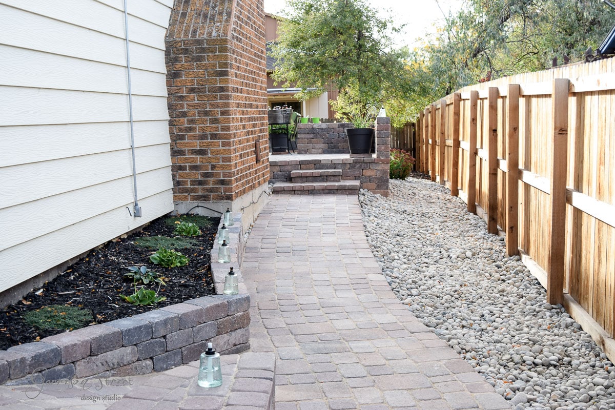 A narrow backyard features a stone path leading to a raised patio with a black planter. On the left, there's a small garden bed with plants and lanterns. A wooden fence borders the right side, with a gravel strip beside it.