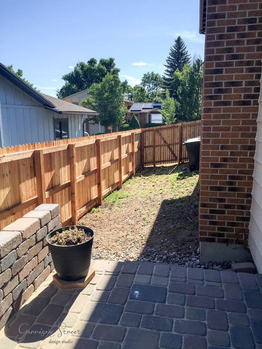 A narrow backyard with a stone patio, wooden fence, and a small planter on the left. The ground is covered with small pebbles and leads to the back gate. The sky is clear with a few clouds.