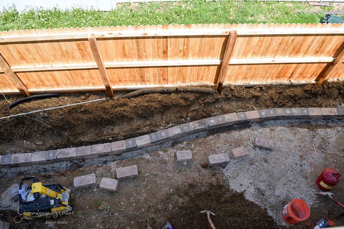 A backyard landscaping project with a partially completed stone border curving along a flower bed. A wooden fence stands in the background. Several construction tools and materials, including bricks and buckets, are scattered around.
