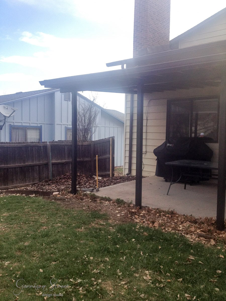 A patio with a wooden pergola attached to a house. There's a black grill and a table on the concrete surface. Fallen leaves are scattered around. A wooden fence separates the yard from neighboring houses. A rake is leaning against the fence.