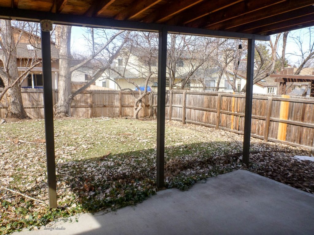 View from a covered patio looking out onto a backyard filled with dry leaves. There are several leafless trees and a wooden fence surrounding the yard. Houses are visible beyond the fence under a clear sky.