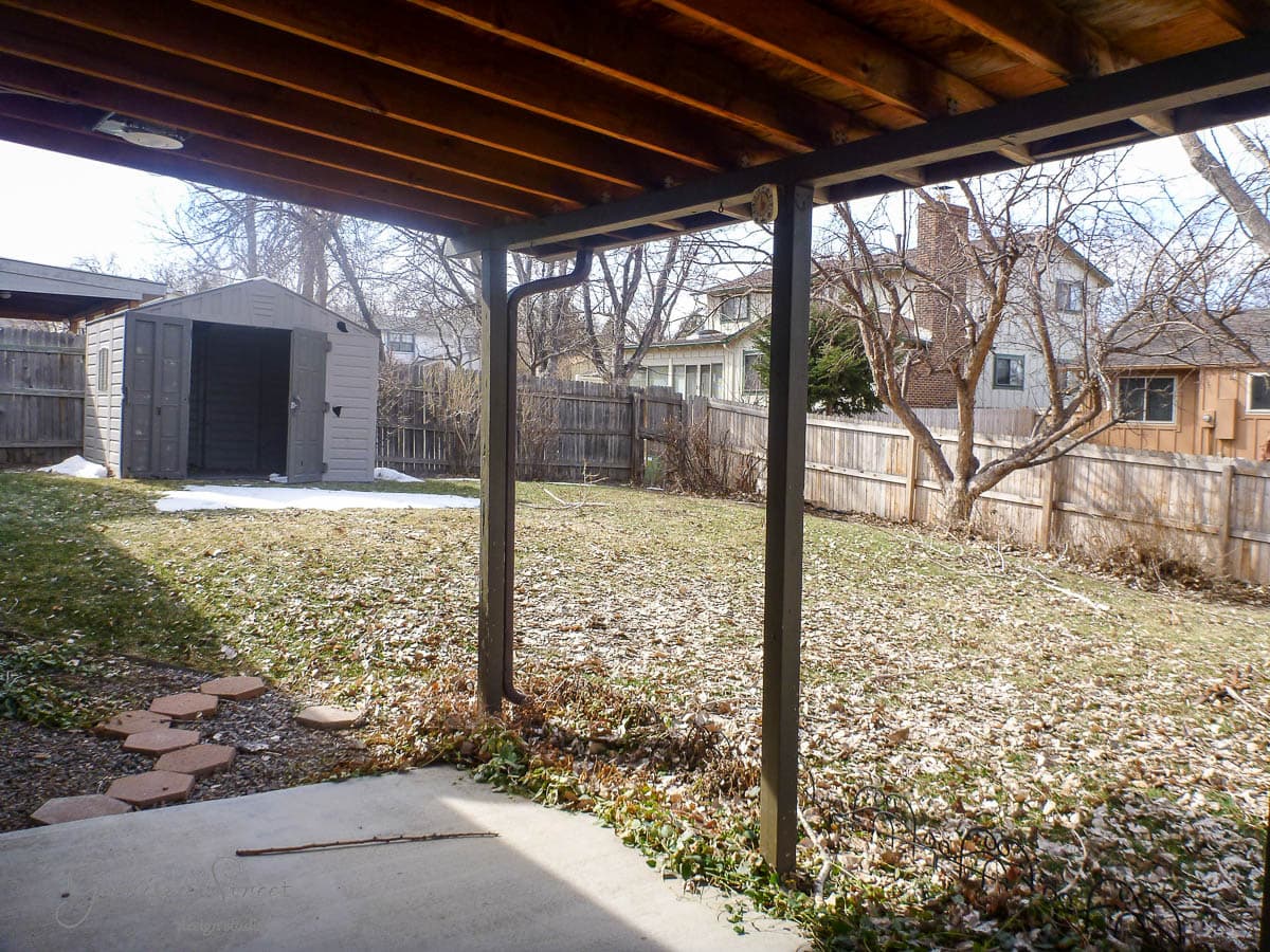 View of a backyard with a wooden pergola, a grassy lawn, and a few patches of snow. A small shed is in the corner, with a wooden fence enclosing the area. Trees are scattered along the fence line, and neighboring houses are visible in the background.