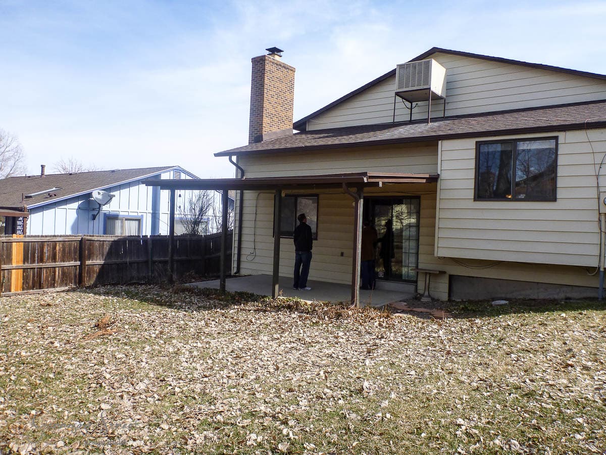 A two-story house with beige siding and a sloped roof. There is a covered patio area with a person standing under it. The backyard has a grass lawn with scattered leaves and is enclosed by a wooden fence. Another house is visible to the left.