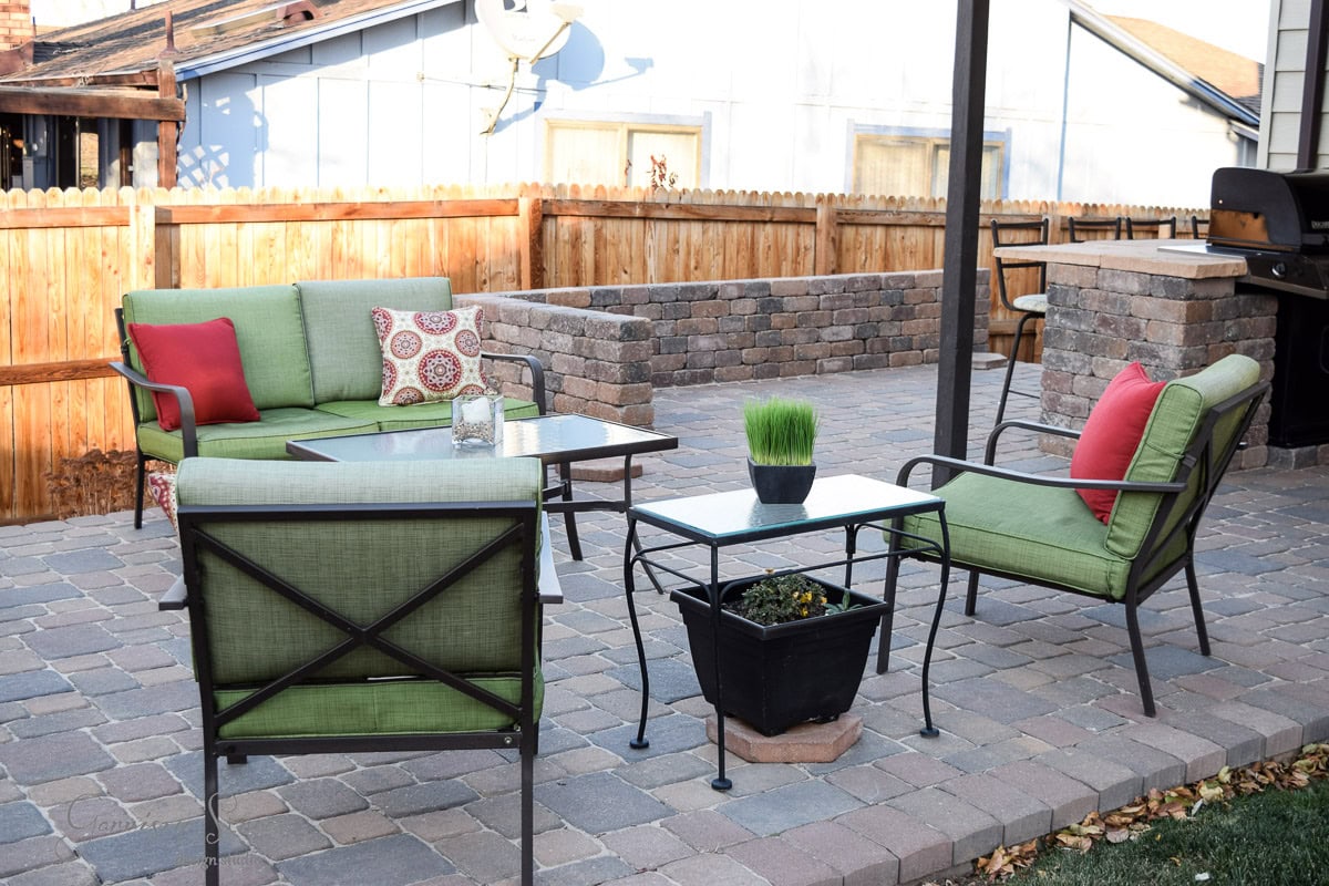 Outdoor patio with green cushioned chairs and a sofa surrounding a glass-top table. Red and patterned pillows add color. A grill and raised brick area are in the background, all set on a stone paver surface, surrounded by a wooden fence.