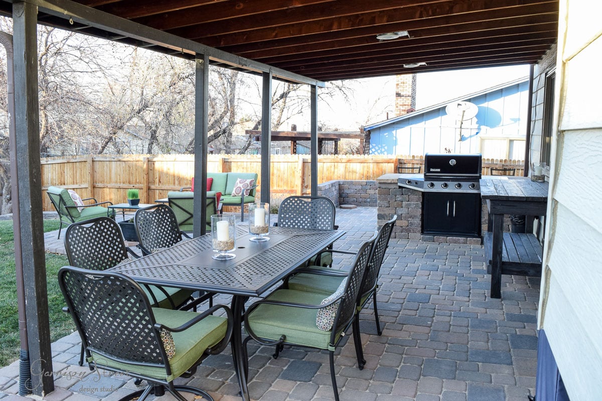 Outdoor patio with a dining table and green cushioned chairs under a wooden pergola. A grill stands nearby. In the background, a seating area with cushions is surrounded by a wooden fence, and trees are visible. Paved stone flooring covers the area.