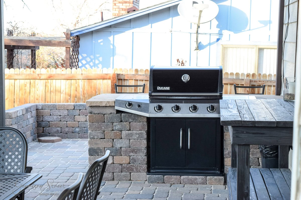 Outdoor patio with a built-in black gas grill set in a stone counter. There's a wooden table nearby and metal chairs with cut-out patterns. A wooden fence and house with blue siding are in the background, with a satellite dish on the roof.