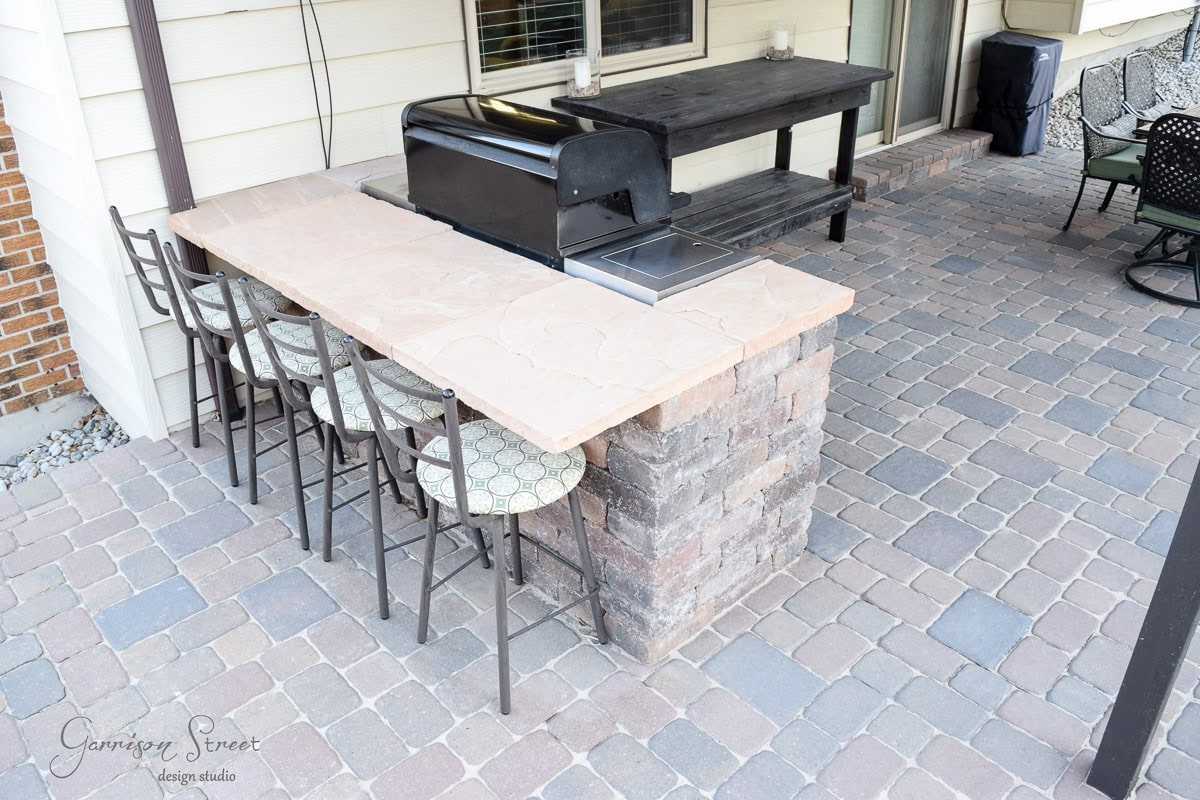 Outdoor kitchen with a stone countertop and built-in grill on a brick patio. Four bar stools with patterned cushions are lined up along the counter, facing the grill. A wooden table is in the background.