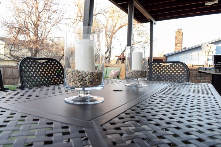 A close-up of a metal outdoor table with black lattice design. Two glass candle holders with white candles and stones inside are placed on it. In the background, there are trees and a chimney, all under a wooden pergola.