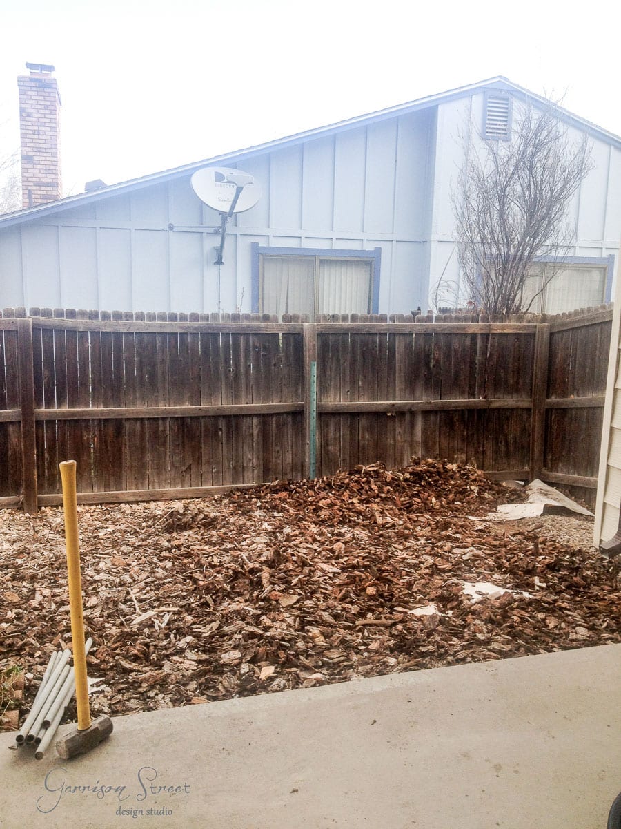 A small backyard with a wooden fence and a backdrop of a house with a chimney and satellite dish. The ground is covered with bark mulch, and there are gardening tools propped against the fence on a concrete area. A leafless shrub is in the corner.