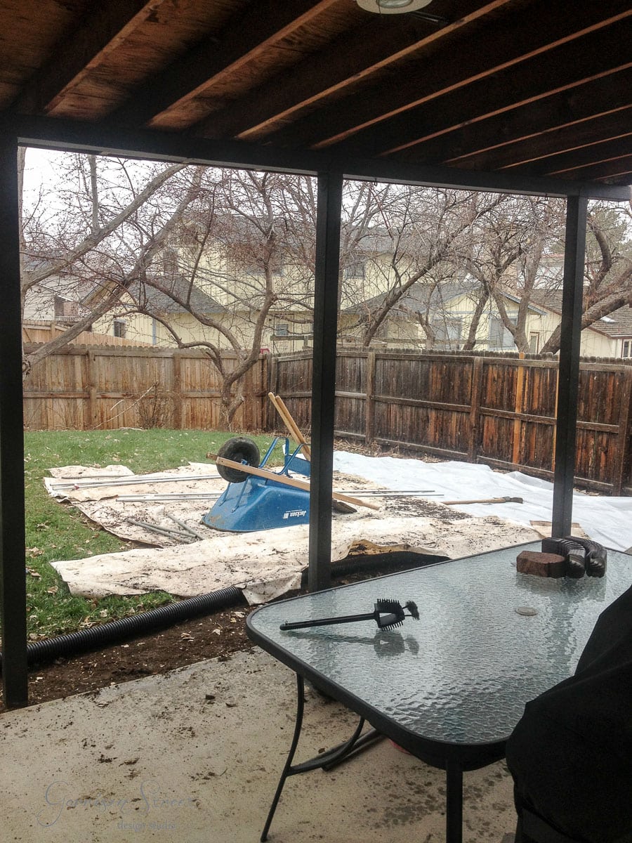 A backyard scene with a glass table in the foreground. Beyond the covered patio, a blue wheelbarrow sits on a tarp-covered area next to bare trees. Wooden fencing surrounds the yard, and it's overcast outside.