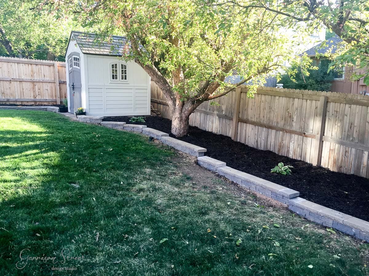A small white shed is nestled under a leafy tree in a backyard. It sits alongside a wooden fence, bordered by a raised garden bed with dark soil. The yard has well-maintained green grass and a gently sloping landscape.