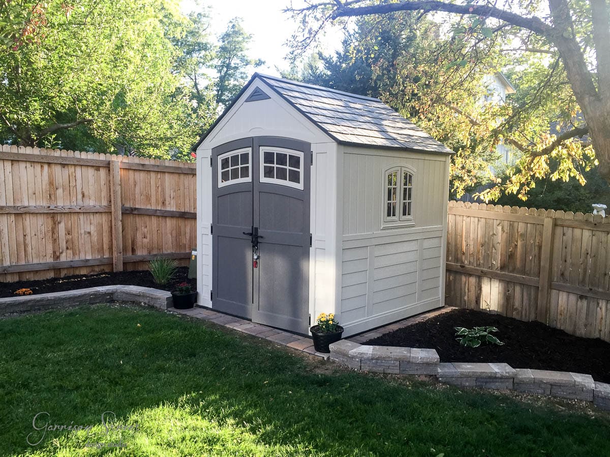 Small garden shed with gray double doors and a window, set on a neatly manicured lawn. It's surrounded by a wooden fence, with trees and shrubs in the background. Patio stones and potted plants decorate the space around the shed.