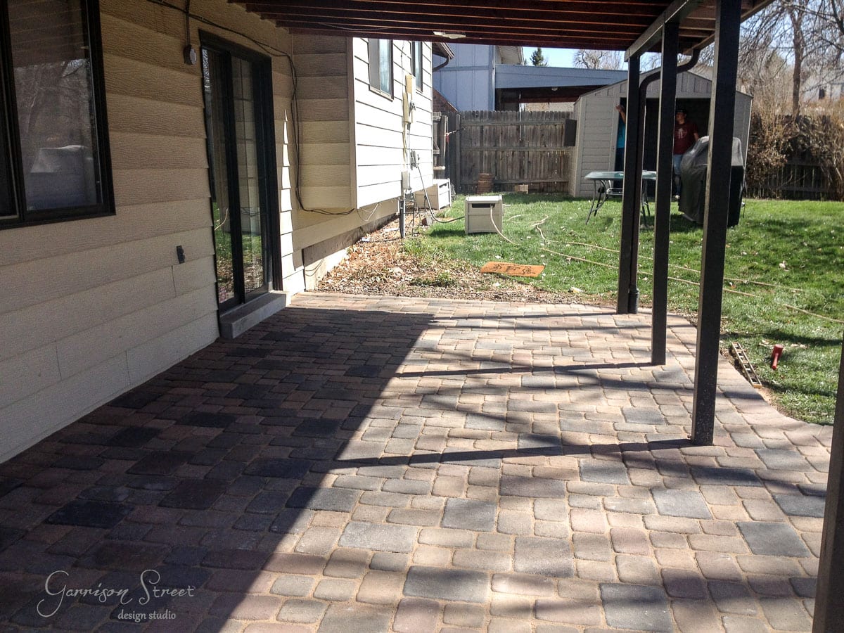 A newly paved patio with interlocking stones extends from a house with sliding glass doors. The area is partly shaded by an overhead structure. In the background, there is a grassy lawn, a wooden fence, and a small shed.