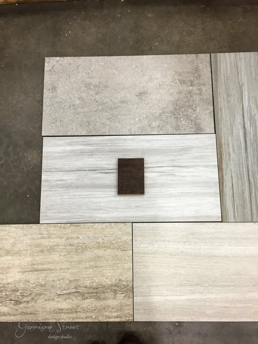 Five rectangular floor tiles of different textures and shades are arranged in a grid pattern. A small, dark brown square tile is placed on top of the center tile. The background is a plain surface.