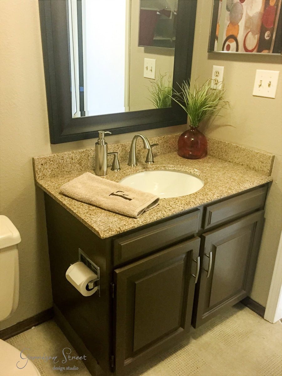 A bathroom vanity with a granite countertop featuring a sink, faucet, and a brown-framed mirror. A beige towel is folded on the counter next to a red vase with green foliage. A toilet and wall art are partially visible.