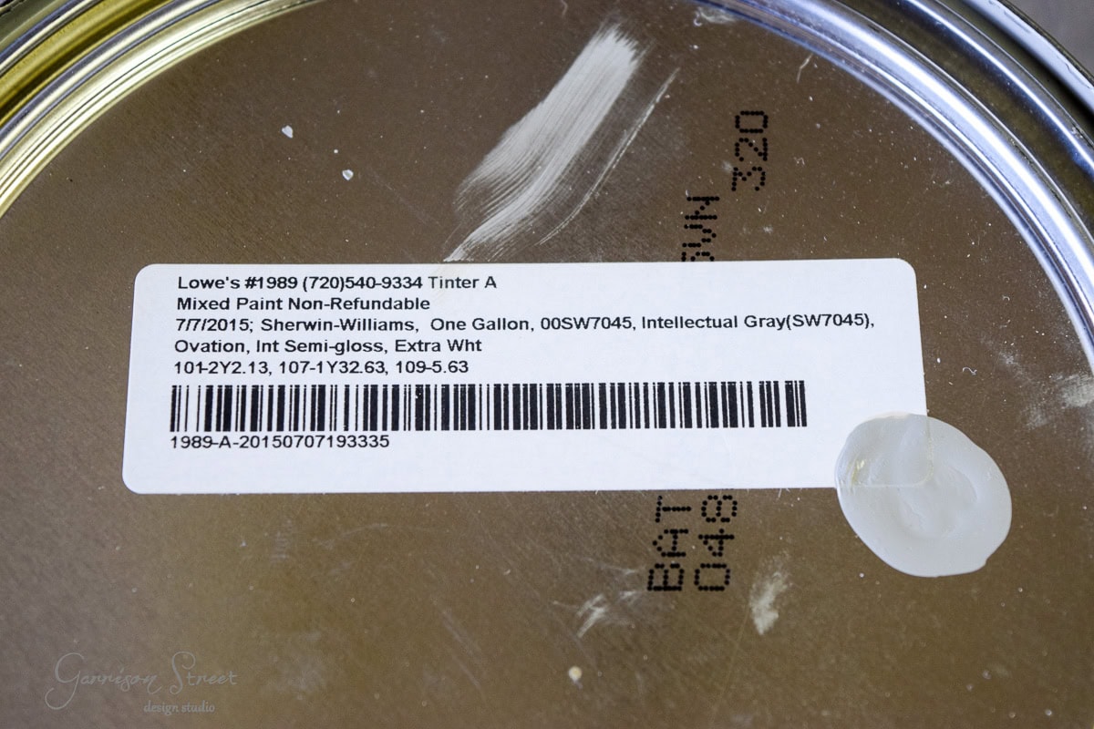 A close-up of a paint can lid with a label. The label reads: "Lowes... Mixed Paint Non-Refundable, Sherwin-Williams, One Gallon, Intellectual Gray (SW7045), Semi-gloss, Extra White." Various codes and measurements are also visible.