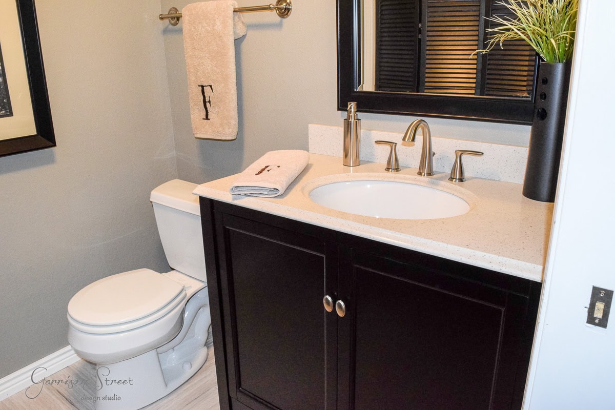 A small bathroom features a white toilet next to a dark wood vanity with a white countertop and silver faucet. A framed picture and towel rack with a beige towel hang on the light gray wall. A vase with grass decorates the counter.