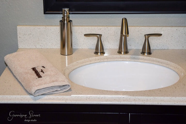 Bathroom sink with a white oval basin set in a speckled countertop. A beige towel with a monogrammed "F" is on the left. Brass faucets include a soap dispenser. A dark framed mirror hangs above, with a logo in the bottom left corner.