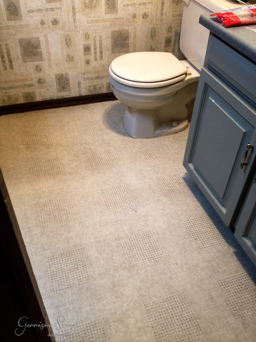 A bathroom with a tan toilet beside a blue cabinet. The floor features a patterned, light-colored vinyl. The walls have a floral wallpaper. A small section of a countertop is visible in the foreground.