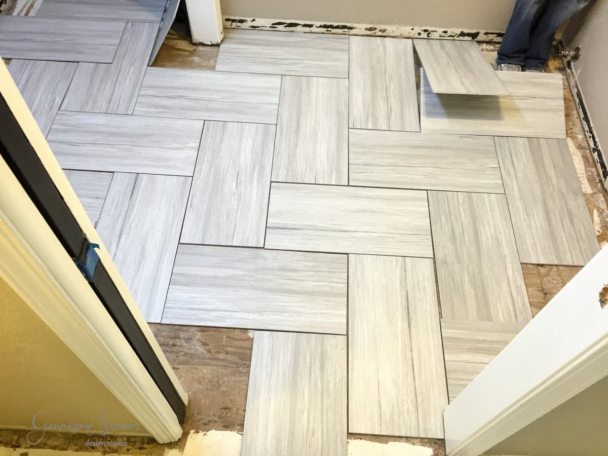 A partially tiled floor in a room under renovation, featuring light gray rectangular tiles in a basket weave pattern. Some tiles are missing, revealing the bare subfloor. The room is bordered by white walls and door frames.