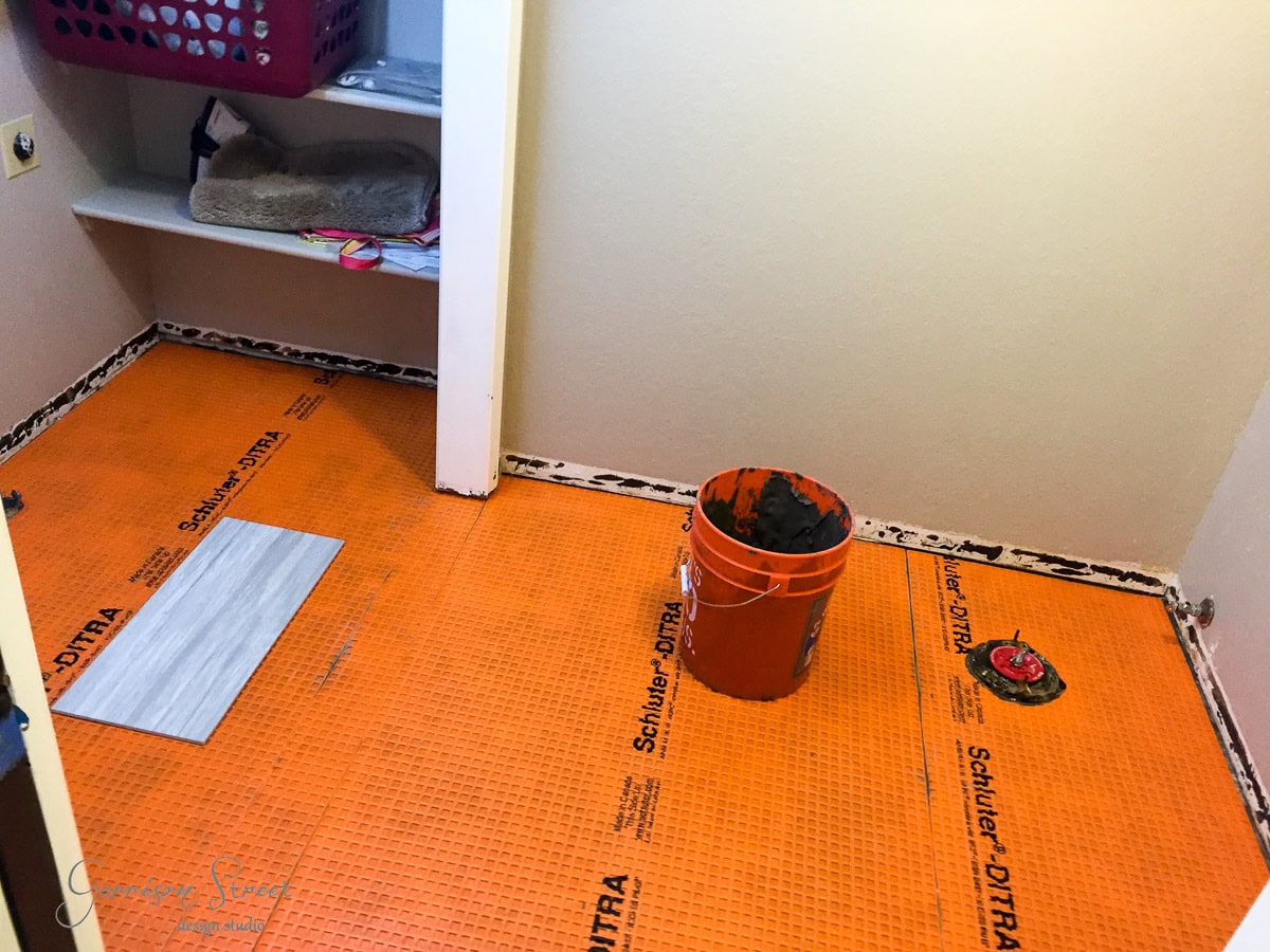 A small room with an orange Schluter DITRA underlayment partially installed on the floor. A single tile is placed for positioning. A red bucket containing mortar and a trowel sits on the floor. A shelf with a basket is in the background.
