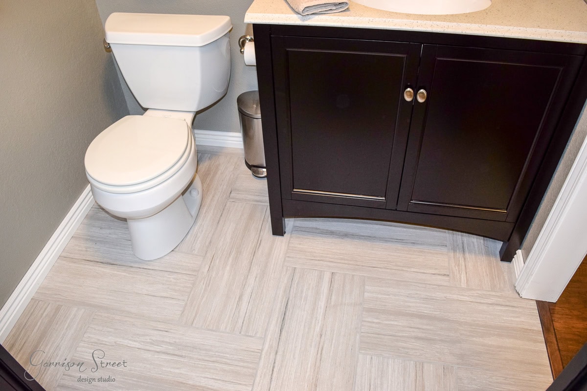 Bathroom with a white toilet and a dark wooden vanity with a light countertop. The floor features large, light gray tiles in a diagonal pattern. A small waste bin sits next to the toilet.