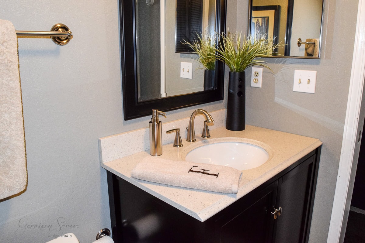 Modern bathroom with a dark wood vanity, white countertop, and chrome faucet. A folded towel with an "F" rests by the sink. Above, a large mirror reflects decorative items, including a tall black vase with green plants.