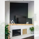 A modern living room features a black accent wall with a mounted TV above a fireplace. A wooden mantel displays decor, and a stack of firewood sits next to the fireplace. White brick and houseplants complete the design.